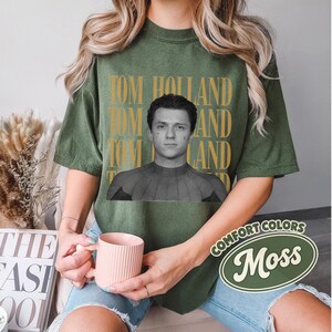 Peut inclure: T-shirt vert olive avec une image en noir et blanc de Tom Holland. Le texte "TOM HOLLAND" est imprim&eacute; en or au-dessus de l'image. Le t-shirt porte une &eacute;tiquette Comfort Colors avec le mot "Moss".