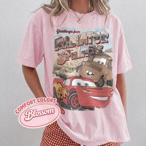 May include: A light pink t-shirt featuring a graphic from the movie Cars. The graphic includes the text "Greetings from Radiator Springs" and images of Lightning McQueen and Mater. The shirt also has the text "Comfort Colors Blossom".