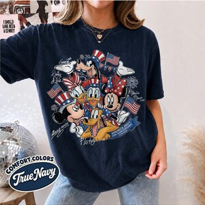 May include: Navy blue t-shirt featuring a graphic of Mickey Mouse, Minnie Mouse, Donald Duck, Daisy Duck, Goofy, and Pluto in patriotic attire. The design includes American flags and the text "America 250th USA Anniversary 1776-2026". The shirt also has the text "Comfort Colors True Navy".