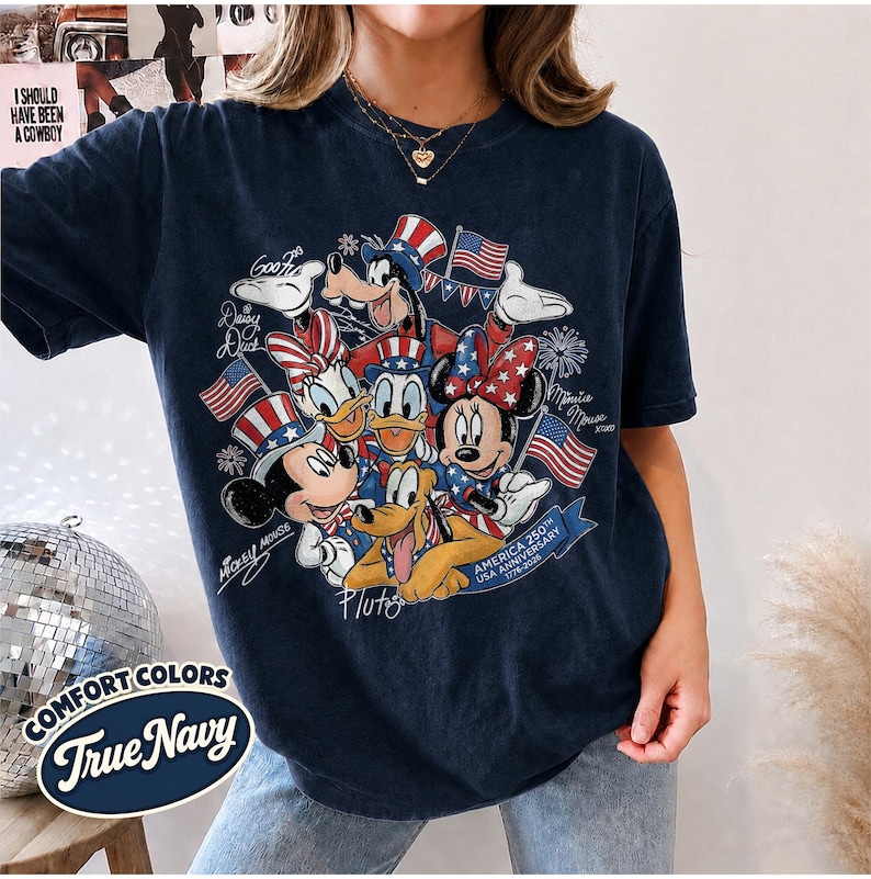 May include: Navy blue t-shirt featuring a graphic of Mickey Mouse, Minnie Mouse, Donald Duck, Daisy Duck, Goofy, and Pluto in patriotic attire. The design includes American flags and the text "America 250th USA Anniversary 1776-2026". The shirt also has the text "Comfort Colors True Navy".