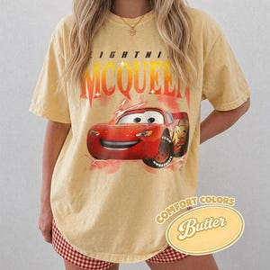 May include: A yellow t-shirt featuring Lightning McQueen from the Cars movie. The graphic includes the text "LIGHTNI MCQUEEN" in red and orange, and the phrase "COMFORT COLORS Butter" in a yellow oval.
