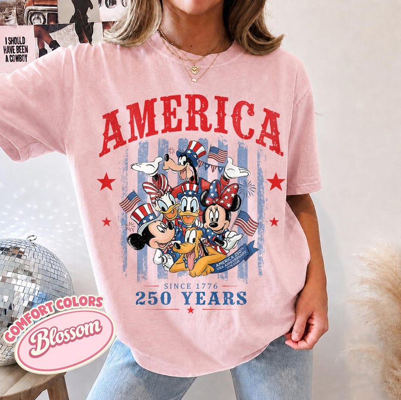 May include: A light pink t-shirt featuring a graphic of Disney characters including Mickey Mouse, Minnie Mouse, Donald Duck, Daisy Duck, Goofy, and Pluto wearing patriotic hats. The text reads AMERICA, SINCE 1776, and 250 YEARS.