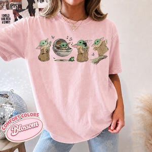 May include: A light pink t-shirt featuring a graphic print of a green creature in various poses, including sitting in a floating pod, drinking from a cup, and standing, accompanied by small musical notes, a frog, and blue butterflies.