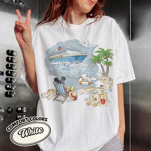 Op de afbeelding: Wit Comfort Colors T-shirt met een strandsc&egrave;ne: cruiseschip, palmbomen, zandkasteel. Mickey Mouse zit in een strandstoel. Het shirt heeft de woorden "Comfort Colors" en "White".