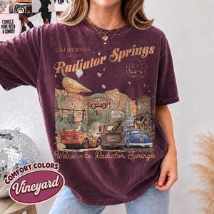 May include: A dark maroon short-sleeve t-shirt featuring a graphic print of Radiator Springs with the text California Est 2012, Radiator Springs, and Welcome to Radiator Springs. The design includes illustrations of three cars, a rock formation, and a water tower.