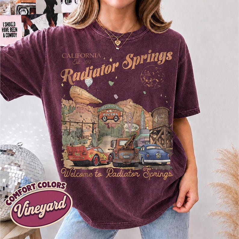 May include: A dark maroon short-sleeve t-shirt featuring a graphic print of Radiator Springs with the text California Est 2012, Radiator Springs, and Welcome to Radiator Springs. The design includes illustrations of three cars, a rock formation, and a water tower.