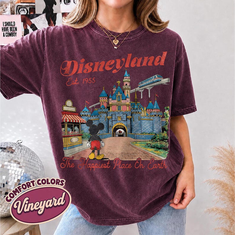 May include: A dark maroon short-sleeve t-shirt featuring a graphic of a blue castle, a monorail, and Mickey Mouse. The text reads Disneyland Est. 1955 and The Happiest Place On Earth. The shirt has a vintage-style wash and a crew neckline.