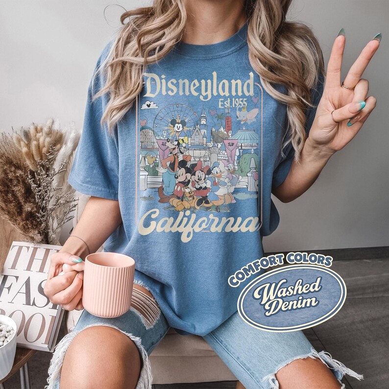 Może przedstawiać: Niebieska koszulka z grafiką Disneyland z Myszką Miki, Myszka Minnie, Kaczorem Donaldem i innymi postaciami. Koszulka ma napisy "Disneyland California" i "Est. 1955". Koszulka jest oznaczona jako "Comfort Colors Washed Denim".