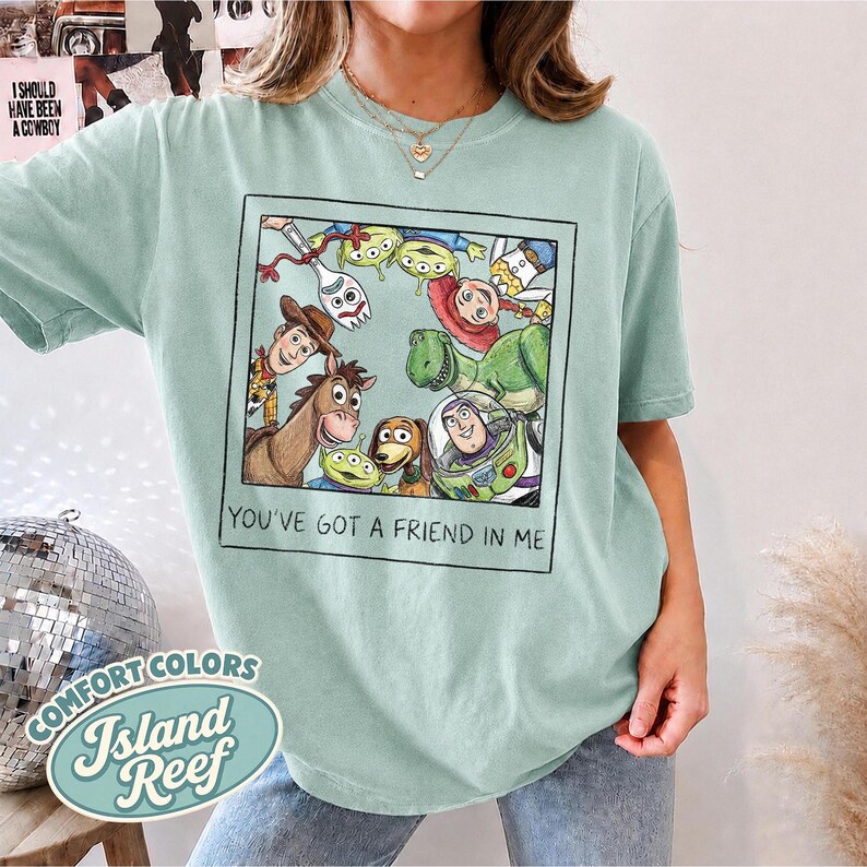 Puede incluir: Una camiseta verde claro con una imagen de dibujos animados enmarcada de personajes de la pel&iacute;cula Toy Story. La imagen incluye a Woody, Buzz Lightyear y otros personajes. El texto "YOU'VE GOT A FRIEND IN ME" est&aacute; debajo.