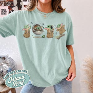 May include: A light green short-sleeve t-shirt featuring a graphic print of a small green creature in various poses, including sitting in a floating pod, standing, and dancing, accompanied by small musical notes and a frog illustration.