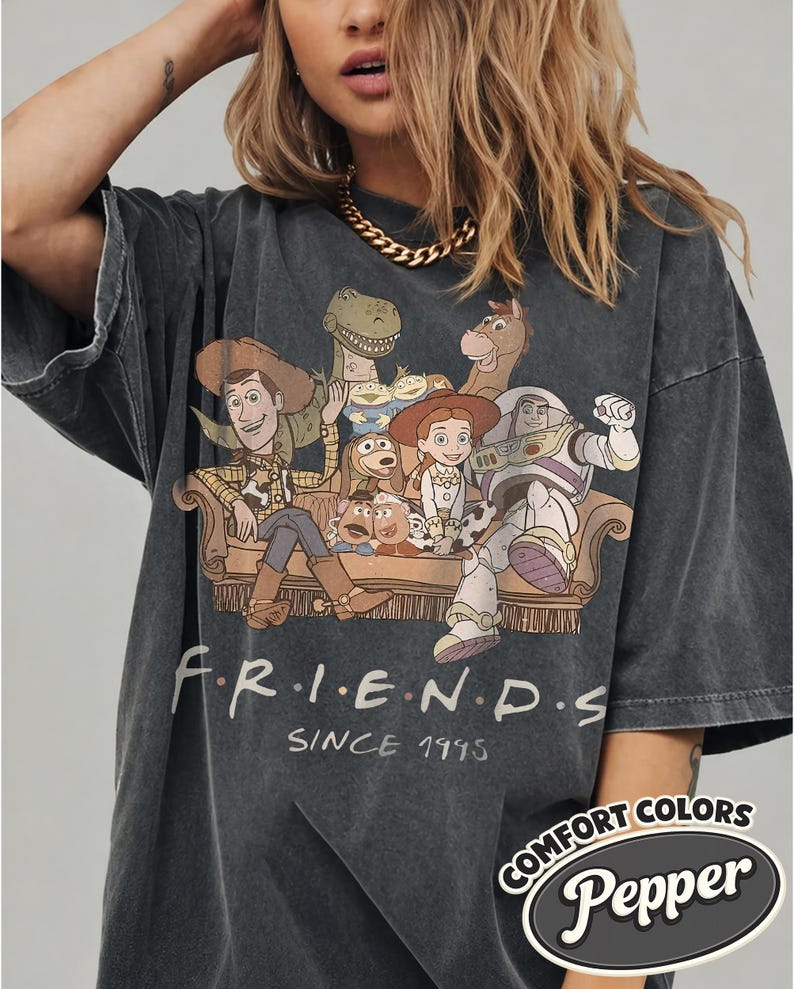 Puede incluir: Camiseta gris oscuro de gran tama&ntilde;o con un gr&aacute;fico de personajes de Toy Story en un sof&aacute;, con la palabra "FRIENDS" y "SINCE 1995" impresas debajo. La camiseta tiene una etiqueta "Comfort Colors Pepper".