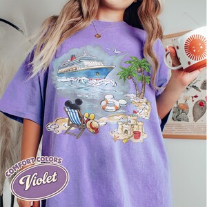 Op de afbeelding: Violet Comfort Colors T-shirt met een strandsc&egrave;ne. Het ontwerp bevat een cruiseschip, palmbomen en een zandkasteel. Mickey Mouse ontspant op een strandstoel. Het shirt heeft ook de tekst "Comfort Colors" en "Violet".