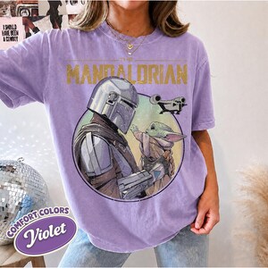 Puede incluir: Una camiseta violeta de manga corta con un estampado gr&aacute;fico de The Mandalorian y Grogu dentro de un marco circular. El texto THE MANDALORIAN est&aacute; impreso en dorado sobre los personajes, con una peque&ntilde;a ilustraci&oacute;n de una nave espacial.