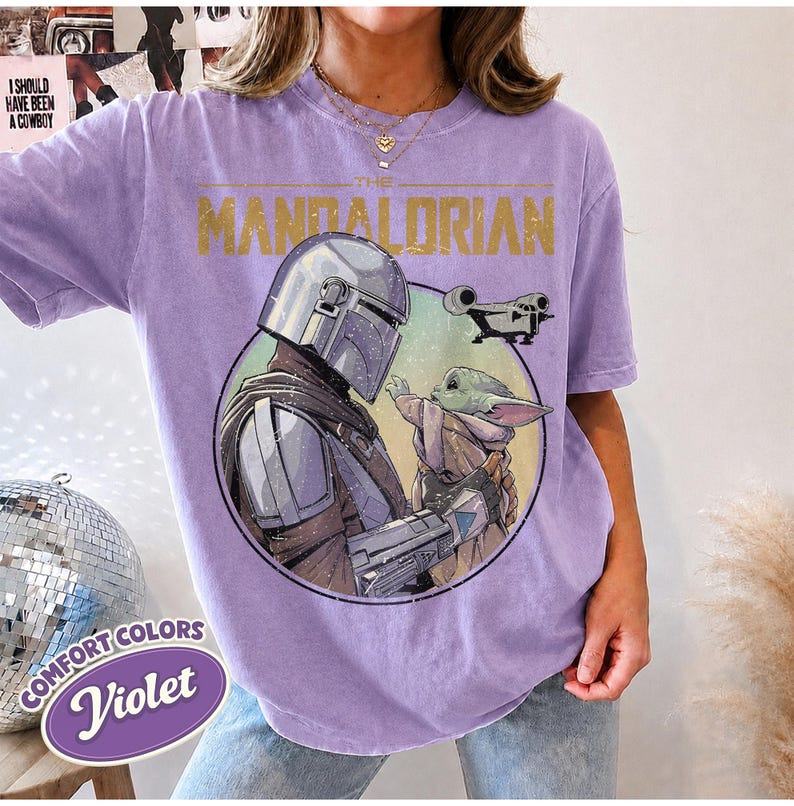 Puede incluir: Una camiseta violeta de manga corta con un estampado gr&aacute;fico de The Mandalorian y Grogu dentro de un marco circular. El texto THE MANDALORIAN est&aacute; impreso en dorado sobre los personajes, con una peque&ntilde;a ilustraci&oacute;n de una nave espacial.