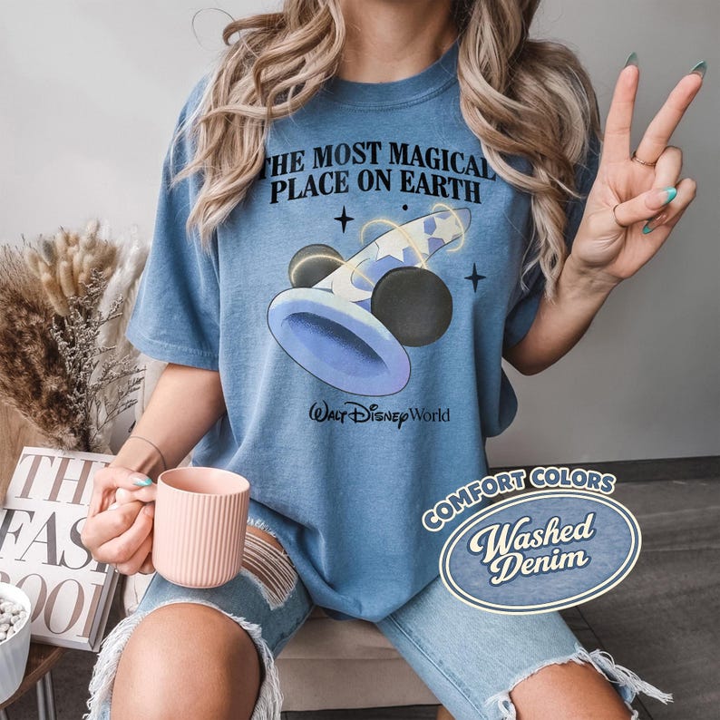 K&ouml;nnte beinhalten: Blaues T-Shirt mit dem Text "THE MOST MAGICAL PLACE ON EARTH" und einer Grafik eines Zauberhuts und Mickey Mouse Ohren. Das Shirt hat auch die Worte "WALT Disney World" und "COMFORT COLORS Washed Denim".