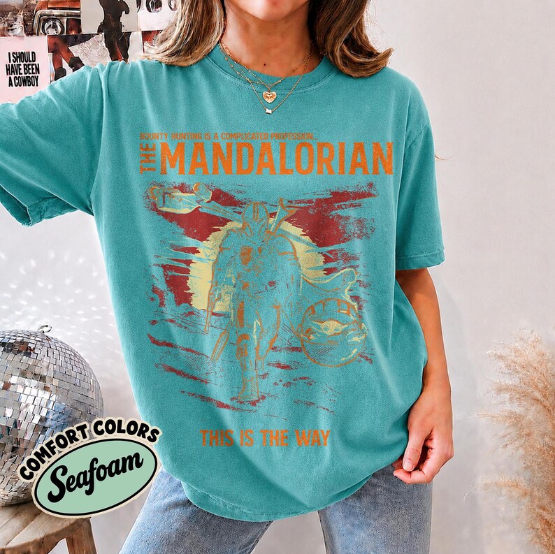 May include: A teal short-sleeve t-shirt featuring a distressed graphic of The Mandalorian with the text "Bounty hunting is a complicated profession," "The Mandalorian," and "This is the way" printed in orange across the chest area.