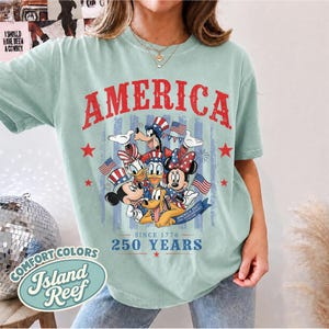 May include: A light green t-shirt featuring a graphic print with the word AMERICA in red letters. The design includes cartoon characters wearing patriotic hats, small American flags, and the text SINCE 1776 250 YEARS in blue lettering.