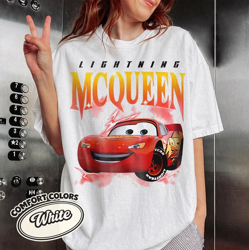 May include: White t-shirt featuring a graphic of Lightning McQueen from the Cars movie. The design includes the text "LIGHTNING MCQUEEN" in yellow and orange, and a red race car. The shirt has a Comfort Colors label.