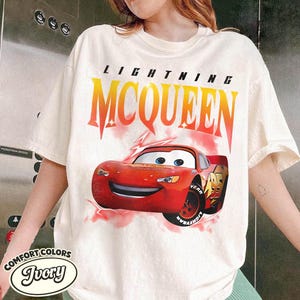 May include: Ivory-colored t-shirt featuring a graphic of Lightning McQueen from the movie Cars. The design includes the text "LIGHTNING MCQUEEN" in a gradient orange and yellow font above the red race car.