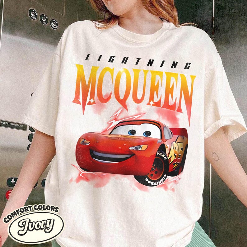 May include: Ivory-colored t-shirt featuring a graphic of Lightning McQueen from the movie Cars. The design includes the text "LIGHTNING MCQUEEN" in a gradient orange and yellow font above the red race car.