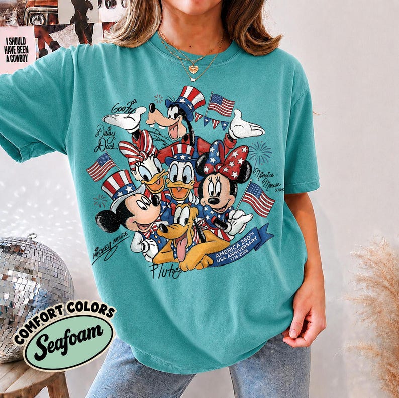 May include: Seafoam green t-shirt featuring a graphic of Disney characters Mickey Mouse, Minnie Mouse, Donald Duck, Daisy Duck, Goofy, and Pluto, with American flags and patriotic hats. The shirt has the text "AMERICA 250TH USA ANNIVERSARY 1776-2026".