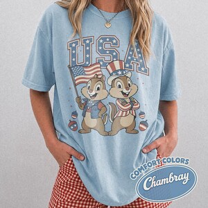 May include: Light blue t-shirt with the word "USA" in red and blue block letters. Features a graphic of two cartoon chipmunks in patriotic attire, holding a flag and wearing a top hat. The shirt has a "Comfort Colors" and "Chambray" logo.
