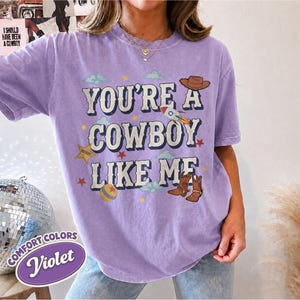 May include: A violet short-sleeve t-shirt featuring the text "YOU'RE A COWBOY LIKE ME" in a bold font. The design includes illustrations of a brown cowboy hat, a rocket ship, a yellow ball, a sheriff star, and brown cowboy boots with stars.