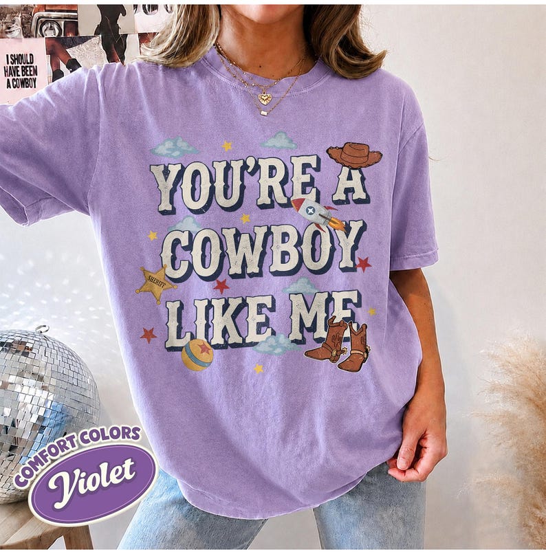 May include: A violet short-sleeve t-shirt featuring the text "YOU'RE A COWBOY LIKE ME" in a bold font. The design includes illustrations of a brown cowboy hat, a rocket ship, a yellow ball, a sheriff star, and brown cowboy boots with stars.