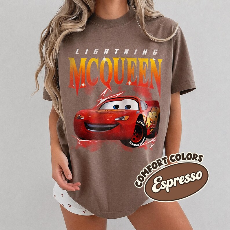 May include: Espresso-colored t-shirt featuring Lightning McQueen from the Cars movie. The design includes the text "LIGHTNING MCQUEEN" in a gradient orange and yellow font above a red cartoon car. The shirt also has the words "COMFORT COLORS Espresso" in a brown oval.