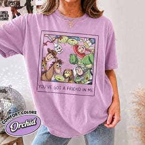 Puede incluir: Camiseta color orqu&iacute;dea con un gr&aacute;fico enmarcado de personajes de Toy Story, incluyendo a Woody y Buzz Lightyear. El texto "YOU'VE GOT A FRIEND IN ME" est&aacute; debajo. La camiseta tambi&eacute;n tiene el texto "COMFORT COLORS" y "Orchid".