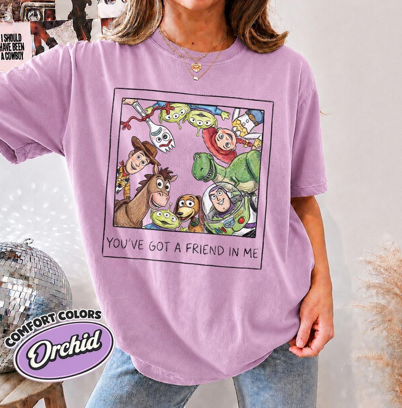 Puede incluir: Camiseta color orqu&iacute;dea con un gr&aacute;fico enmarcado de personajes de Toy Story, incluyendo a Woody y Buzz Lightyear. El texto "YOU'VE GOT A FRIEND IN ME" est&aacute; debajo. La camiseta tambi&eacute;n tiene el texto "COMFORT COLORS" y "Orchid".
