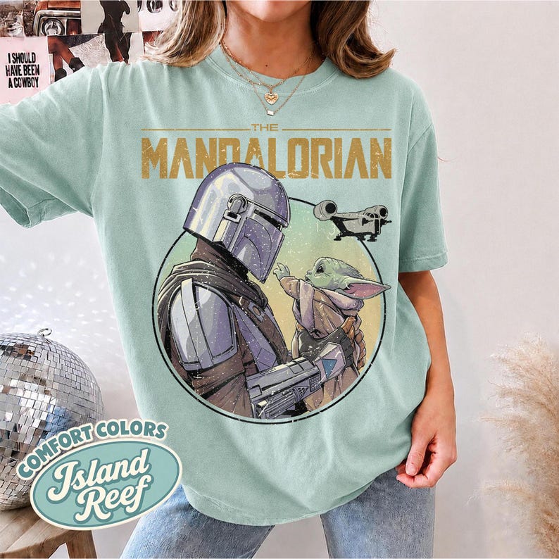 Puede incluir: Una camiseta de color verde claro con un estampado gr&aacute;fico de The Mandalorian y Grogu dentro de un marco circular. El texto "THE MANDALORIAN" est&aacute; impreso en dorado sobre los personajes, con una peque&ntilde;a ilustraci&oacute;n de una nave espacial.