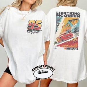 May include: White t-shirt featuring Lightning McQueen from the Cars movie. The front displays "95" with "Lightning McQueen" text. The back showcases a racing scene with Lightning McQueen and the text "Lightning McQueen Racing Series".