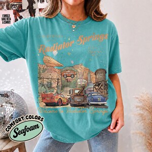 May include: A teal short-sleeve t-shirt featuring a graphic print of Radiator Springs with the text "Radiator Springs" and "Welcome to Radiator Springs." The illustration includes a red race car, a brown tow truck, and a blue car in a desert landscape.