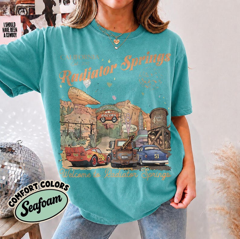 May include: A teal short-sleeve t-shirt featuring a graphic print of Radiator Springs with the text "Radiator Springs" and "Welcome to Radiator Springs." The illustration includes a red race car, a brown tow truck, and a blue car in a desert landscape.