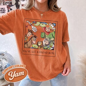 Puede incluir: Camiseta naranja Comfort Colors con un gr&aacute;fico enmarcado de personajes de Toy Story. La frase "YOU'VE GOT A FRIEND IN ME" est&aacute; impresa debajo del gr&aacute;fico. La camiseta tambi&eacute;n tiene los logotipos de Comfort Colors y Yam.