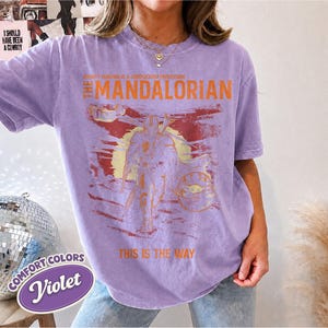 May include: A light purple short-sleeve t-shirt featuring a graphic print of The Mandalorian and Grogu. The text reads: BOUNTY HUNTING IS A COMPLICATED PROFESSION, THE MANDALORIAN, and THIS IS THE WAY. The design includes a sun and landscape.