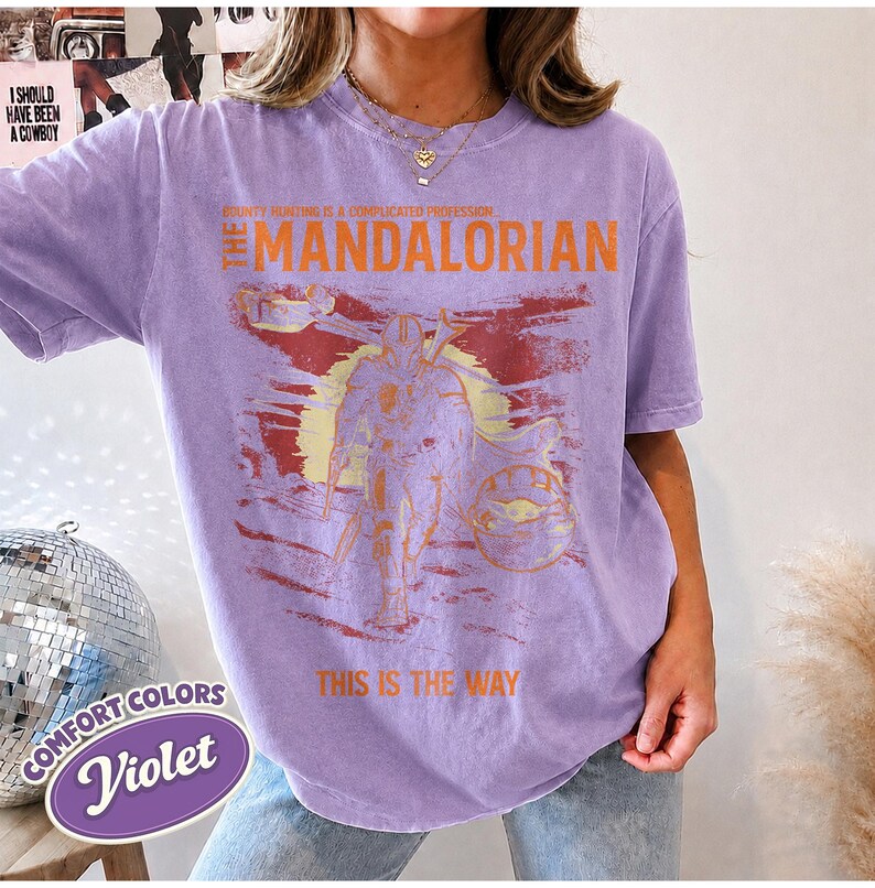 May include: A light purple short-sleeve t-shirt featuring a graphic print of The Mandalorian and Grogu. The text reads: BOUNTY HUNTING IS A COMPLICATED PROFESSION, THE MANDALORIAN, and THIS IS THE WAY. The design includes a sun and landscape.