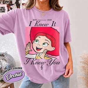 May include: An orchid-colored short-sleeve t-shirt featuring a graphic of a cartoon character with red hair and a red hat. The text reads EST 2026, I Knew It, and I Knew You with a small musical note symbol centered below the graphic design.