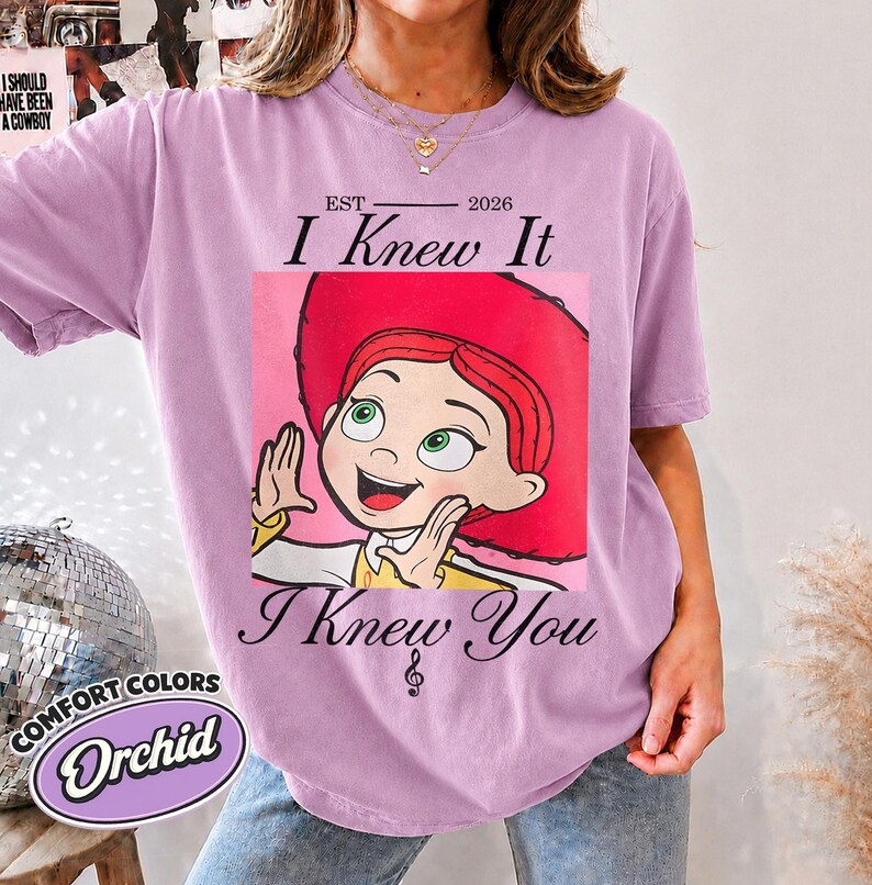 May include: An orchid-colored short-sleeve t-shirt featuring a graphic of a cartoon character with red hair and a red hat. The text reads EST 2026, I Knew It, and I Knew You with a small musical note symbol centered below the graphic design.