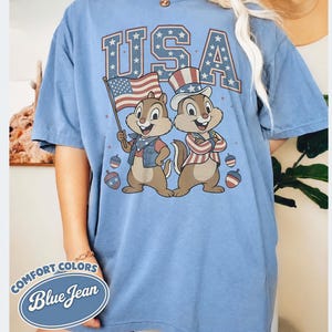 May include: A light blue Comfort Colors t-shirt with a graphic of two cartoon chipmunks in patriotic attire, the word "USA" in a star-filled font, and the text "Blue Jean". The chipmunks are holding a flag and wearing a top hat.