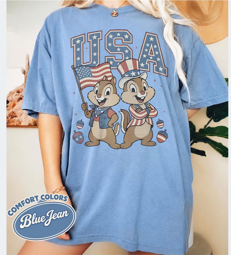 May include: A light blue Comfort Colors t-shirt with a graphic of two cartoon chipmunks in patriotic attire, the word "USA" in a star-filled font, and the text "Blue Jean". The chipmunks are holding a flag and wearing a top hat.