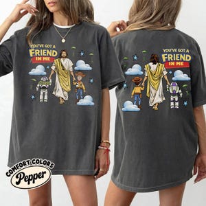 May include: Dark grey t-shirt featuring the text "YOU'VE GOT A FRIEND IN ME" above a graphic of Jesus, Woody, and Buzz Lightyear. The shirt has a crew neck and short sleeves. The brand name "COMFORT COLORS Pepper" is printed on the bottom left.