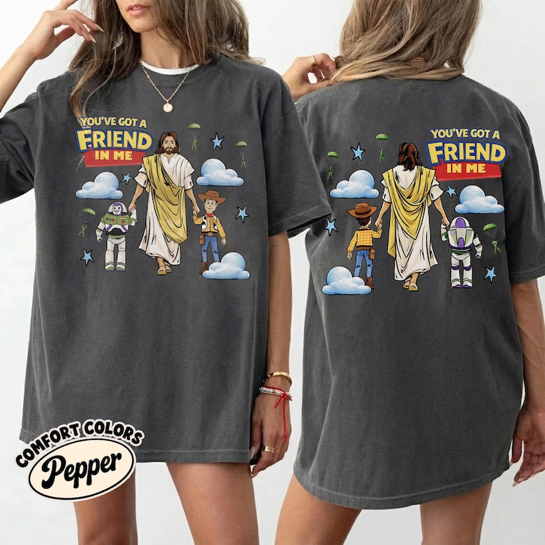 May include: Dark grey t-shirt featuring the text "YOU'VE GOT A FRIEND IN ME" above a graphic of Jesus, Woody, and Buzz Lightyear. The shirt has a crew neck and short sleeves. The brand name "COMFORT COLORS Pepper" is printed on the bottom left.