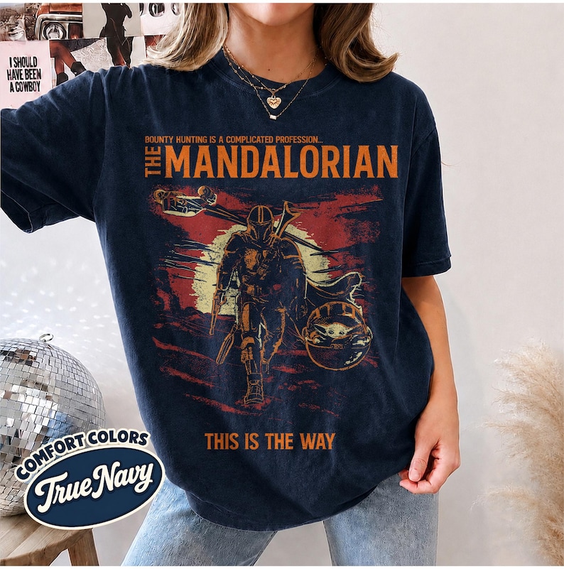 May include: A dark navy blue short-sleeve t-shirt featuring a graphic print of The Mandalorian. The design includes the text "BOUNTY HUNTING IS A COMPLICATED PROFESSION... THE MANDALORIAN" and "THIS IS THE WAY" in orange, with a distressed illustration.