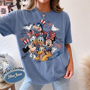 May include: Blue jean colored t-shirt featuring a graphic of Disney characters in patriotic attire, including Mickey and Minnie Mouse, Donald and Daisy Duck, Goofy, and Pluto. The shirt commemorates the USA's 250th anniversary.