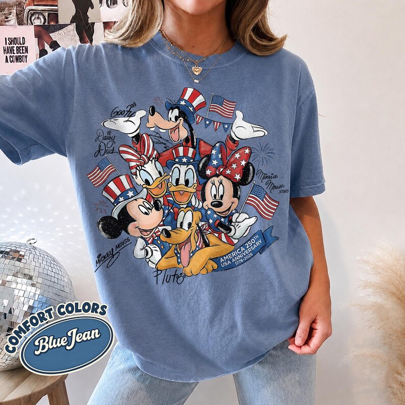May include: Blue jean colored t-shirt featuring a graphic of Disney characters in patriotic attire, including Mickey and Minnie Mouse, Donald and Daisy Duck, Goofy, and Pluto. The shirt commemorates the USA's 250th anniversary.