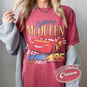 K&ouml;nnte beinhalten: Karmesinrotes T-Shirt mit Lightning McQueen aus dem Film Cars. Das Design zeigt das Bild der Figur, den Text "Lightning McQueen" und "Think Fast 95". Das Shirt hat einen Vintage-Look mit einem Comfort Colors-Label.