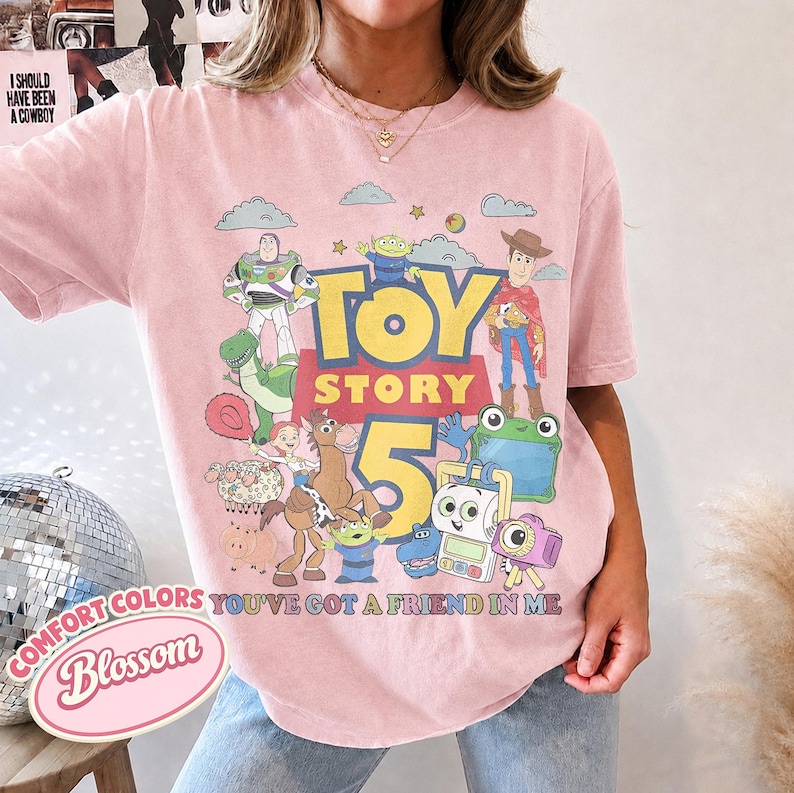 May include: A light pink t-shirt featuring a graphic print of Toy Story characters including Buzz Lightyear, Woody, Jessie, and Bullseye. The text reads Toy Story 5 and You've Got A Friend In Me. The shirt has a crew neckline and short sleeves.