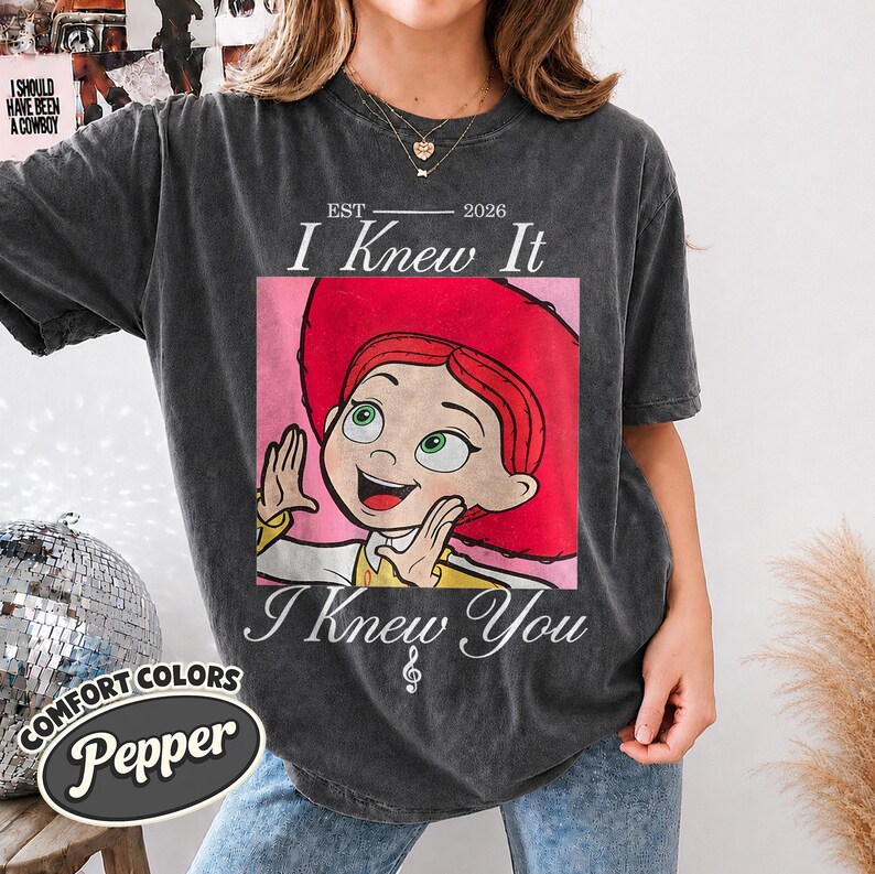May include: A charcoal gray short-sleeve t-shirt featuring a graphic of a red-haired character in a red hat. The text reads EST 2026 I Knew It I Knew You with a small musical note symbol. The shirt has a vintage washed appearance.